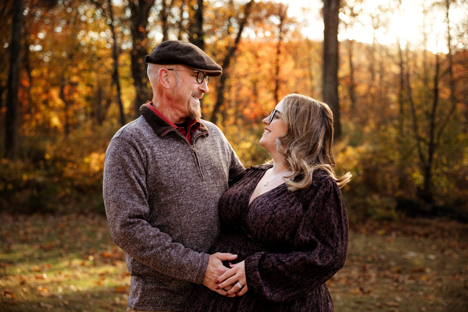 Photo of a soon to be mom and dad looking at one another in front of the fall trees.