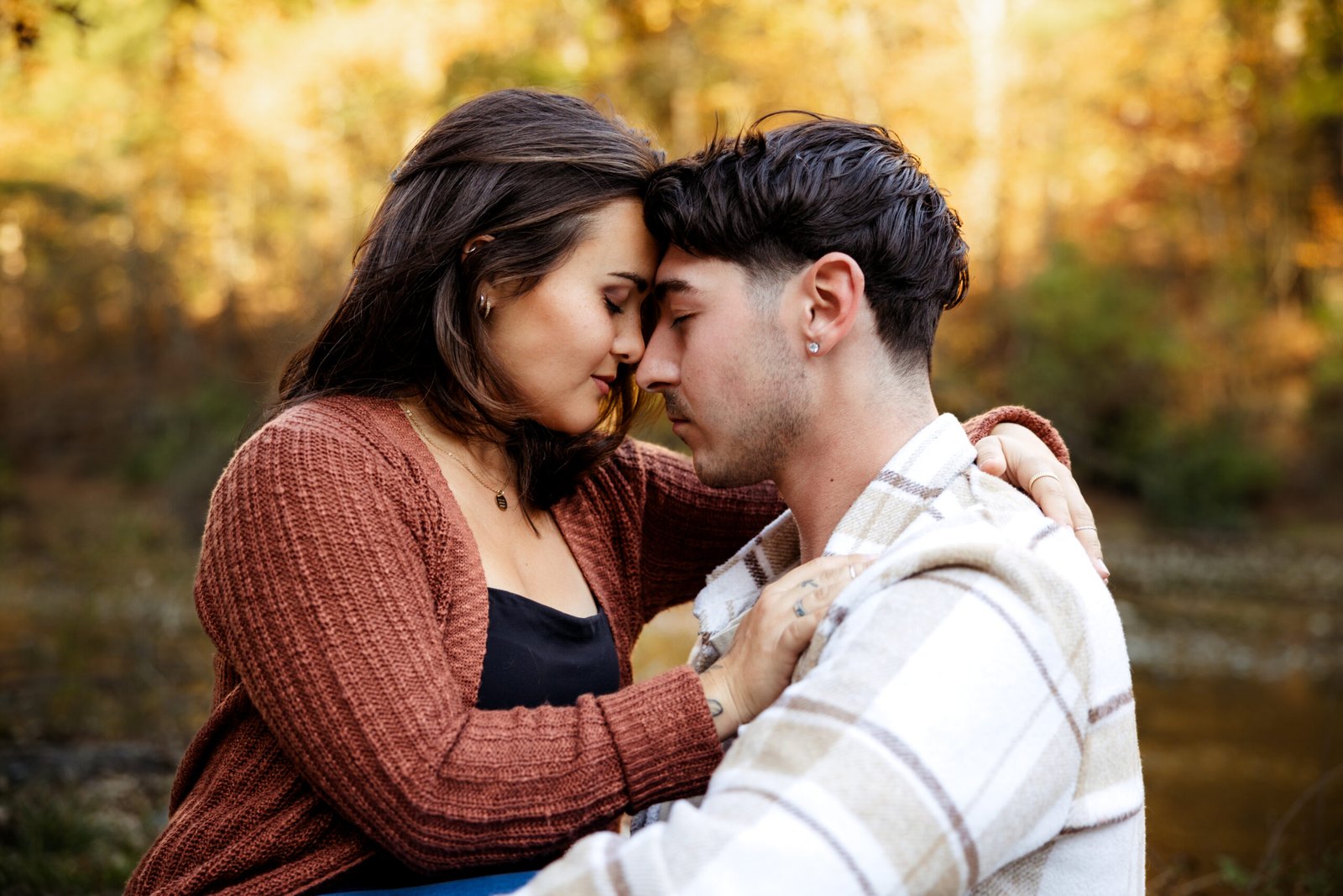 Couple sitting in front of body of water with their heads resting against one another.