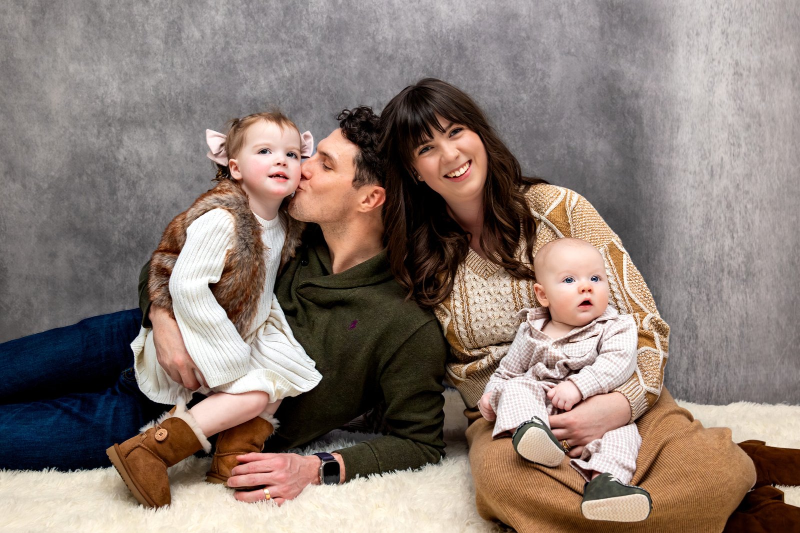 Family of four sitting together on the floor with Dad giving his daughter a kiss on her cheek.